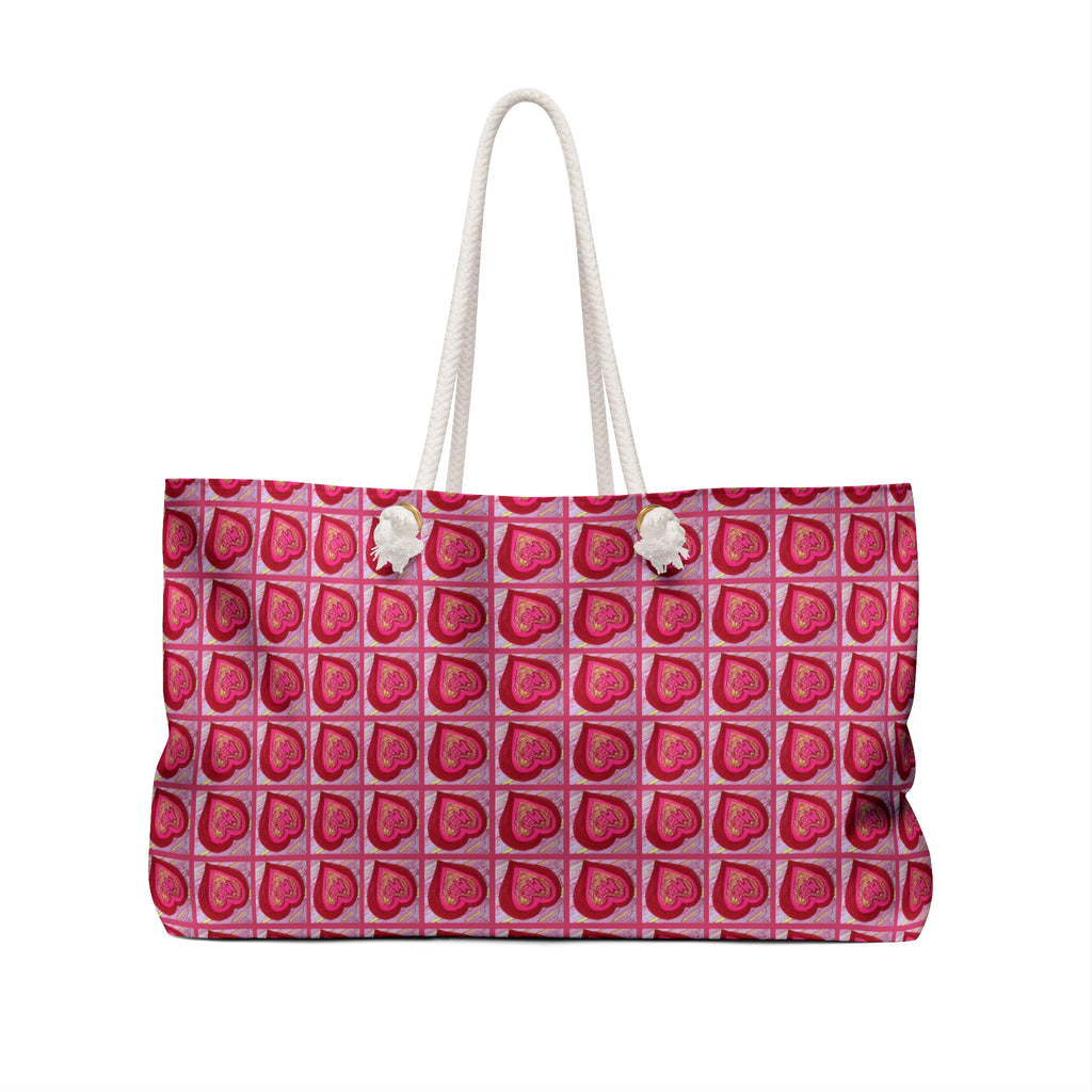 A tote bag with a repeating pattern of pink and red hearts on a light pink background, featuring white rope handles with white pom-pom accents, shown against a plain white background.